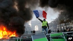 Seorang demonstran mengibarkan bendera Perancis di dekat barikade yang dibakar di jalan Champs-Elysees dengan latar belakang, Arc de Triomphe saat berlangsungnya unjuk rasa menentang kenaikan pajak bahan bakar, di Paris, Perancis, 24 November 2018. (Foto: dok).