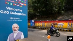 在印度新德里，一名摩托车骑手驶过描绘印度自由斗争的墙上涂鸦，左侧是欢迎G20峰会各国代表的海报。(2023年9月7日)