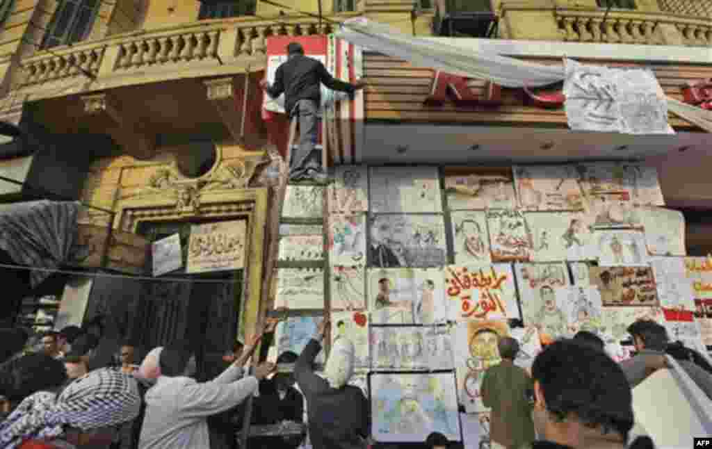 An artists hangs another political drawing on the shop front of a closed fast food outlet, in a street which has become a meeting point and workshop for artists to create such posters, at the continuing anti-government demonstration in Tahrir Square in do