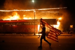 A protester carries a U.S. flag upside down, a sign of distress, next to a burning building, May 28, 2020, in Minneapolis.