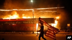 A protester carries a U.S. flag upside down, a sign of distress, next to a burning building, May 28, 2020, in Minneapolis. 
