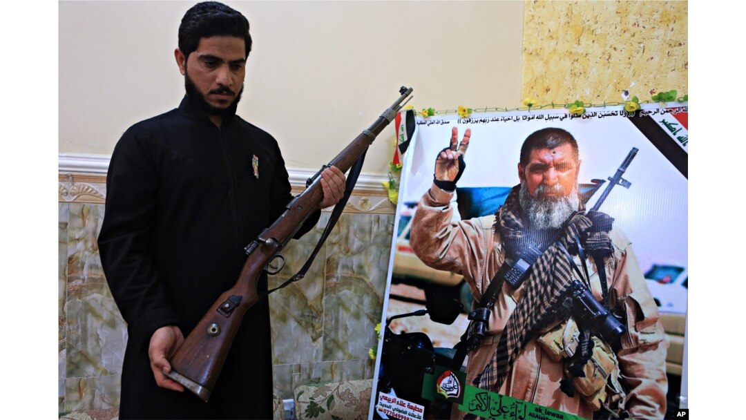 FILE - Talib, the son of Iraqi sniper Ali Jayad al-Salhi, 65, a volunteer with Popular Mobilization Forces killed fighting Islamic State militants, poses with a weapon next to his father's poster in his home in Basra, Iraq, Dec. 5, 2017.