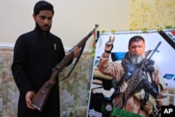 FILE - Talib, the son of Iraqi sniper Ali Jayad al-Salhi, 65, a volunteer with Popular Mobilization Forces killed fighting Islamic State militants, poses with a weapon next to his father's poster in his home in Basra, Iraq, Dec. 5, 2017.