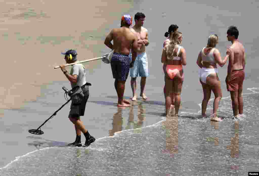 Un hombre peina la arena con un detector de metales, mientras la gente acude en masa a la playa para disfrutar del fin de semana largo.