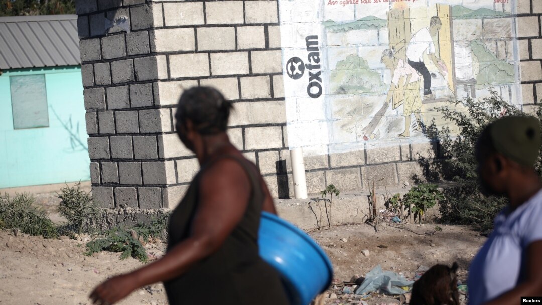 Dua perempuan berjalan melewati sebuah tembok dengan tulisan Oxfam di dindingnya di Port-au-Prince, Haiti (foto: ilustrasi). 
