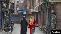 FILE - A Kashmir girl rides her bike past Indian security force personnel standing guard in front of closed shops on a street in Srinagar, Oct. 30, 2019.