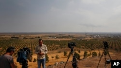 FILE - TV journalists work on a hilltop in Ceylanpinar, Sanliurfa province, southeastern Turkey, as in the background smoke billows from a fire in Ras al-Ayn, Syria, Oct. 20, 2019. 