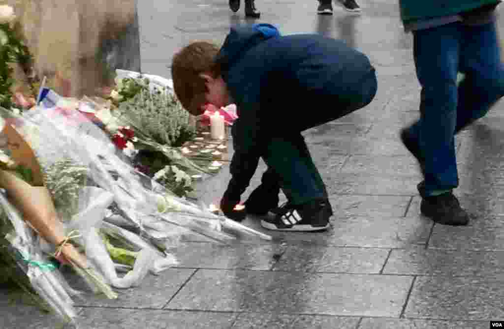 Seorang anak laki-laki menyalakan lilin di depan plakat yang menyatakan penghormatan kepada 15 orang yang tewas dalam serangan maut di bagian utara Paris tahun lalu (13/11). (VOA/L. Bryant)