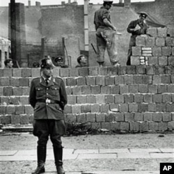 Một cảnh vệ Tây Berlin đứng trước bức tường bê tông chia cắt Đông và Tây Berlin ở Bernauer Strasse, trong khi các công nhân Đông Berlin chất các khối gạch lên bức tường để xây cao bức tường chắn này, 7/10/1961.