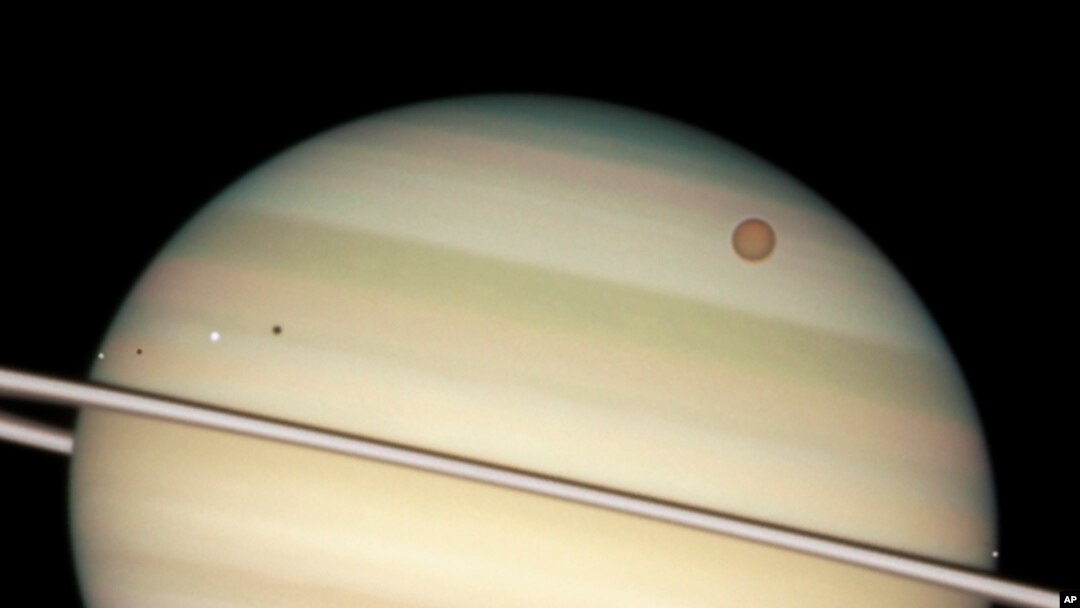 FILE - This image provided by NASA captures the transit of several moons across the face of Saturn. The giant orange moon Titan – larger than the planet Mercury – can be seen at upper right.