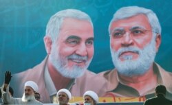 Iraqi clerics look on as they stand near a banner depicting senior Iranian military commander General Qassem Soleimani and Iraqi militia commander Abu Mahdi al-Muhandis, Jan. 3, 2021.