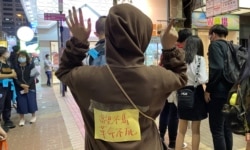 香港市民阿澧11月30日晚在旺角街站寫心意卡給12港人，她在背上貼上標語，高舉反送中運動”五大訴求、缺一不可”的手勢。(美國之音特約記者湯惠芸攝)