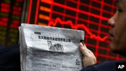 Seorang pria memegang surat kabar sambil mengamati papan elektronik yang menampilkan harga-harga saham di lantai bursa di Beijing (5/12). (AP/Andy Wong)
