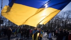 Seorang perempuan mengibarkan bendera Ukraina raksasa dalam protes memperingati dua tahun invasi besar-besaran Rusia, di Gerbang Brandenburg, Berlin, Sabtu, 24 Februari 2024. (Foto: AP)