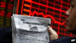 Seorang pria memegang surat kabar sambil mengamati papan elektronik yang menampilkan harga-harga saham di lantai bursa di Beijing (5/12). (AP/Andy Wong)