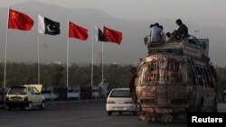 People sit on top of a bus as they go past flags of Pakistan and China that are displayed along a road.