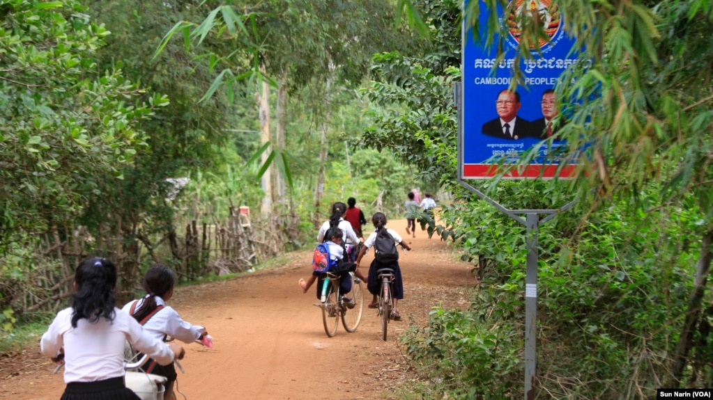 Children ride their bicycles past a sign bearing the Cambodian People's Party logo and the faces of Prime Minister Hun Sen and National Assembly President Heng Samrin in Banteay Meas village, Taches commune, Kampong Chhnang province, Nov. 8, 2017. 