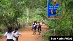 Children ride their bicycles past a sign bearing the Cambodian People's Party logo and the faces of Prime Minister Hun Sen and National Assembly President Heng Samrin in Banteay Meas village, Taches commune, Kampong Chhnang province, Nov. 8, 2017. 