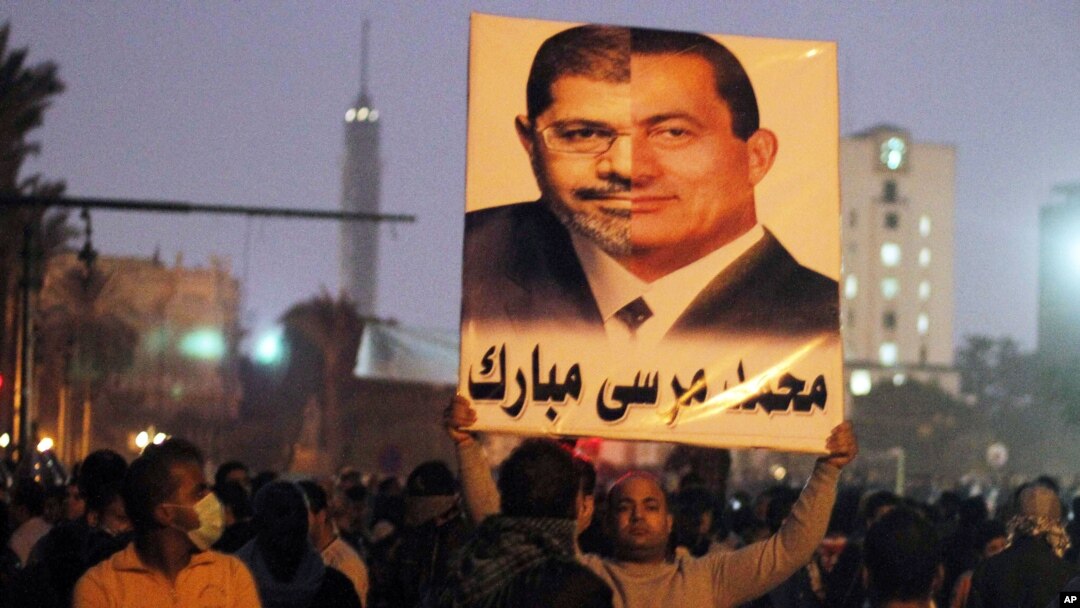 Một người biểu tình phản đối tại Quảng trường Tahrir, ở Cairo, Ai Cập, cầm bức ảnh ráp nối của Tổng thống đương quyền Morsi (phần trái) và cựu Tổng thống Mubarak. Công chúng tức giận vì ông Morsi và nhóm Huynh đệ Hồi giáo của ông nắm quá nhiều quyền hành