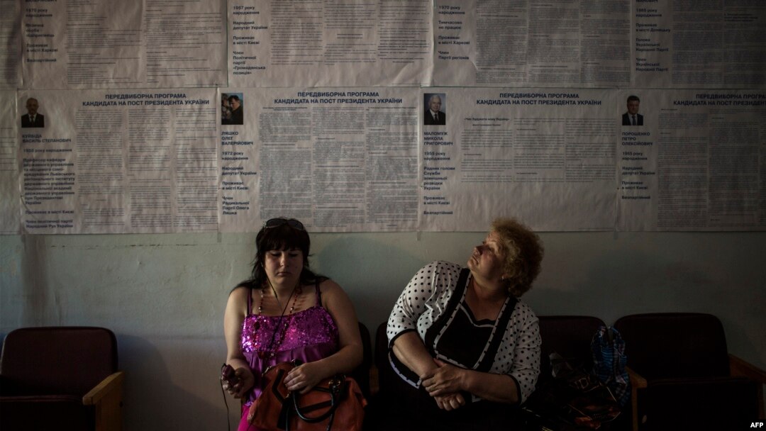 Dua orang perempuan duduk dekat poster pemilu di tempat pemungutan suara di Krasnoarmiisk, provinsi Donetsk, Ukraina timur (24/5).