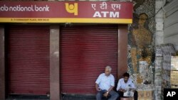 A security guard, right, reads a newspaper as he sits in front of a closed ATM in New Delhi, India, April 18, 2018. Several ATMs were seen to have run dry across the country, reportedly from a surge in demand of cash withdrawals. 