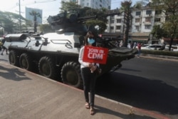 An anti-coup protester holding a sign reading "Join in CDM (Civil Disobedience Movement)" poses for a photo in front of an armored personnel carrier deployed outside the Central Bank of Myanmar building, in Yangon, Myanmar, Feb. 15, 2021.