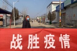A man wearing a face mask stands near a banner reading "defeat the epidemic" stretched across the entrance to Donggouhe village in northern China's Hebei province, Jan. 29, 2020.