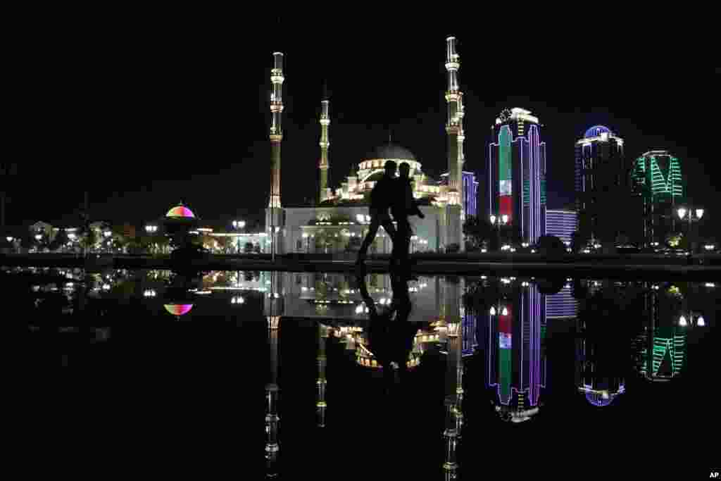 Dua orang berjalan saat malam hari di pusat kota Grozny, ibukota Chechnya, di Rusia selatan, sementara bayangan bangunan Masjid Besar Grozny tercermin di dalam air kolam.