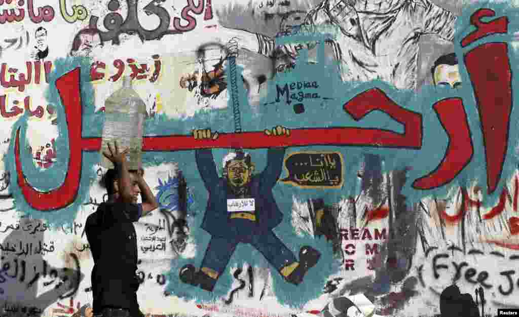 Un hombre transportando un contenedor de agua sobre su cabeza pasa frente a un muro cubierto de graffitis en oposición al depuesto ex presidente Mohamed Morsi con el mensaje &quot;Fuera&quot;, en la plaza Tahrir (Liberación), de El Cairo.