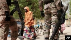 In this photo taken Wednesday, April 8, 2015, a woman walks past Nigerian Soldiers at a checkpoint in Gwoza, Nigeria, a town newly liberated from Boko Haram. Each day brings new reports of atrocities, with mass graves being discovered in towns seized ba