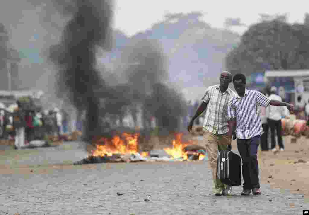 Two men carry a suitcase past a burning barricade in Bujumbura, Burundi, after the government issued and ordered all university campuses to close down. Bujumbura has been hit by street protests since Sunday as security forces confront demonstrators who say a third term for President Pierre Nkurunziza would violate the country&#39;s constitution.