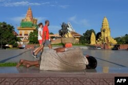 កុមារអនាថាម្នាក់ដេកនៅក្នុងសួនកម្សាន្តមួយកន្លែង ខណៈភ្ញៀវទេសចរពីរនាក់ដើរកាត់ នៅកណ្តាលក្រុងរាជធានីភ្នំពេញ ថ្ងៃទី ២៣ ខែមិថុនា ឆ្នាំ២០២០។