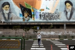 In this Friday, April 3, 2020 photo, a man crosses an empty street under portraits of the late Iranian revolutionary founder Ayatollah Khomeini, right, and Supreme Leader Ayatollah Ali Khamenei, left, in Tehran, Iran.