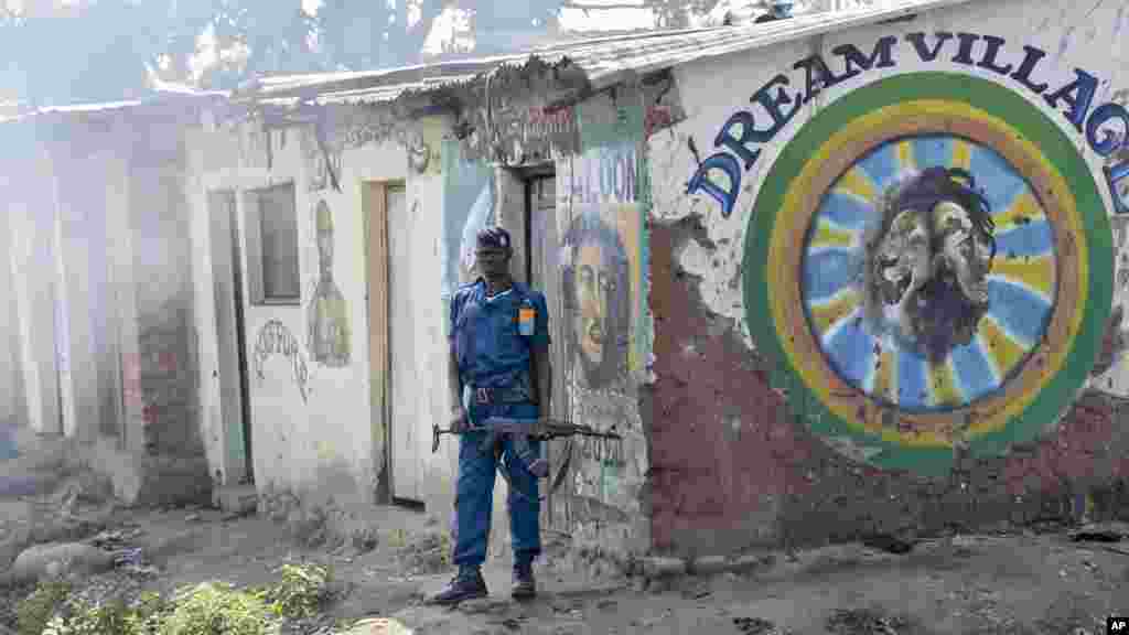Um policial do Burundi guardando posto à porta da barbearia "Dream Village". A fumaça que se aproxima dele é de gás lacrimogéneo, (esquerda) durante os confrontos entre manifestantes da oposição e forças de segurança no distrito de Cibitoke da capital Bujumbura, Burundi, 29 de Maio, 2015.
