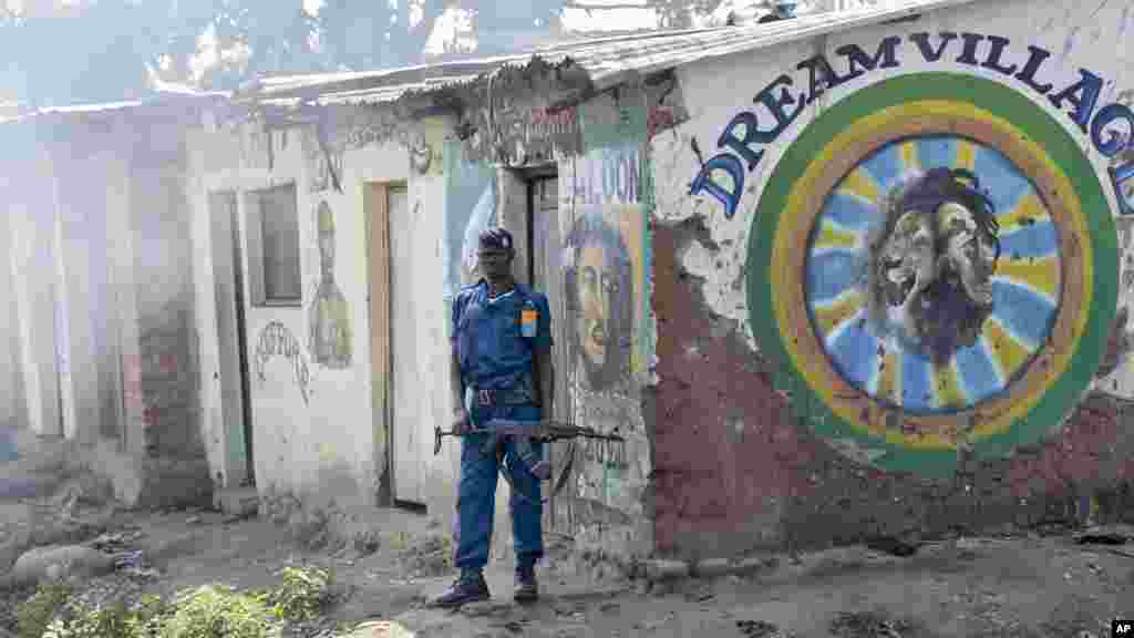 Um policial do Burundi guardando posto à porta da barbearia &quot;Dream Village&quot;. A fumaça que se aproxima dele é de gás lacrimogéneo, (esquerda) durante os confrontos entre manifestantes da oposição e forças de segurança no distrito de Cibitoke da capital Bujumbura, Burundi, 29 de Maio, 2015.