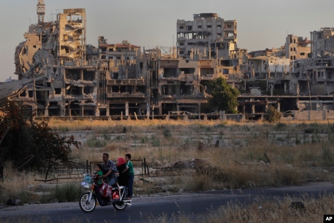 FILE - People ride their motorcycle by damaged buildings in the old town of Homs, Syria, Aug. 15, 2018.