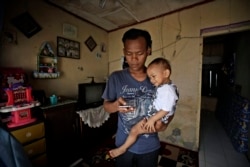 In this Monday, April 20, 2020, photo, Budi Santosa, who was a cook at a Chinese restaurant before losing his job amid the coronavirus outbreak, checks his phone as he carries his young son in his rented house in Jakarta, Indonesia. (AP Photo/Dita Alangka