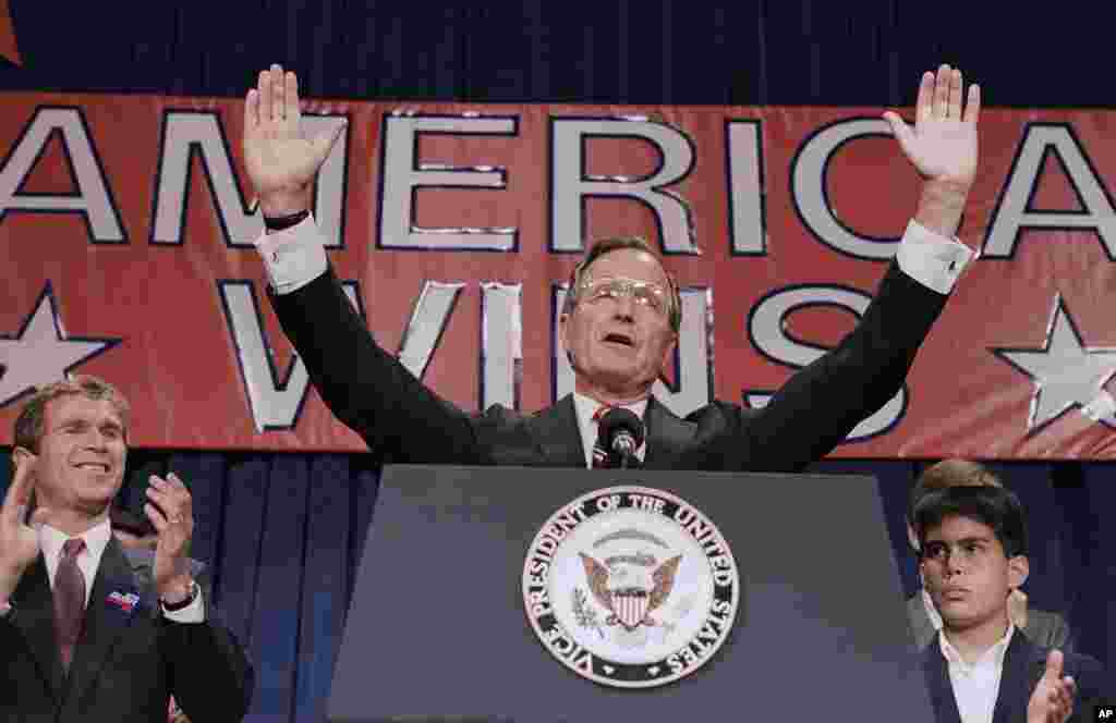 This November 9, 1988, photo shows President-elect George H. W. Bush holding his hands up to acknowledge the crowds applause, and ask them to allow him to continue his speech during his victory rally with grandson, George P. Bush, right, and son, George W. Bush.