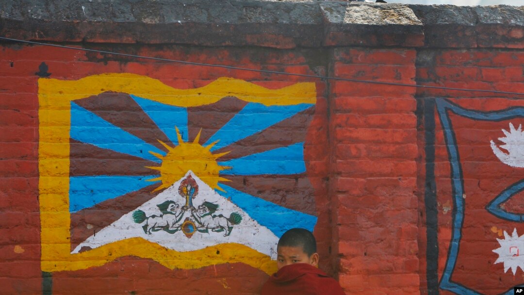 Seorang biksu Tibet di pengasingan berdiri di depan dinding yang dicat dengan bendera Tibet di Katmandu. (Foto: AP)