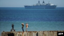 FILE - People take pictures on a remain of the World War II "Mulberry" artificial harbour as a warship is anchored in the English Channel in Arromanches-les-Bains, Normandy.