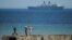 FILE - People take pictures on a remain of the World War II "Mulberry" artificial harbour as a warship is anchored in the English Channel in Arromanches-les-Bains, Normandy.