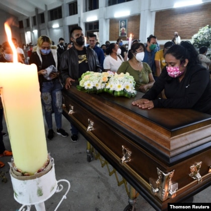 A group of people gather for nighttime Mass behind a coffin. A woman places her hands on top of the coffin.