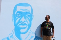 Ruhel Islam, owner of Gandhi Mahal Restaurant, poses next to a mural of George Floyd located across the street from the ruins of the restaurant. Despite losing his restaurant, Islam continues to support the Black Lives Matter movement. (K. Khan/VOA)
