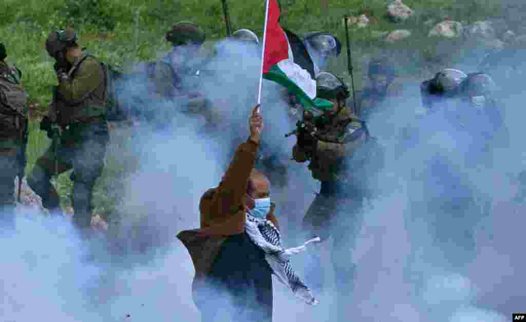 A Palestinian protester raises a national flag as a member of the Israeli forces points his gun, amid smoke from tear gas, during a demonstration against the establishment of Israeli outposts on their lands, in Beit Dajan, in the occupied West Bank.