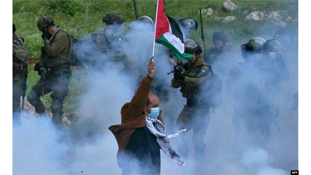 Seorang pria lansia Palestina mengibarkan bendera nasional ketika seorang anggota pasukan Israel menodongkan senjatanya, di tengah asap gas air mata, selama aksi protes menentang pengambilalihan tanah Palestina di Beit Dajan, Tepi Barat (foto: dok).