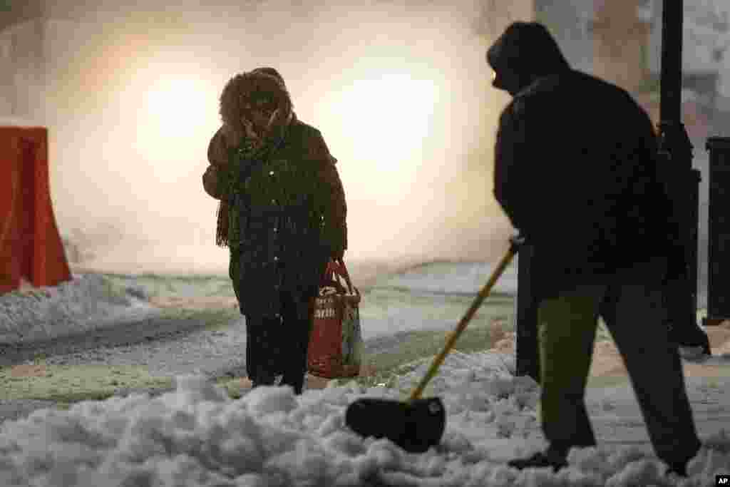 Seorang perempuan berjalan di trotoar sementara pekerja membersihkan trotoar itu dari salju akibat badai musim dingin di Philadelphia (14/3). (AP/Matt Rourke)