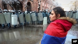 زنی با پرچم ارمنستان در جریان تجمعی در ایروان برای تحت فشار قرار دادن نیکول پاشینیان، نخست وزیر ارمنستان، برای استعفا به دلیل توافق صلح با جمهوری آذربایجان، مقابل نیروهای پلیس ضد شورش ایستاده است. (عکس آرشیوی از آذر ۱۳۹۹) 