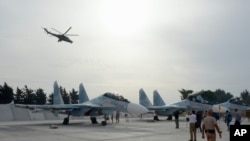 ເຮືອບິນລົບອາຍພົ່ນ Su-30 ທີ່ຈອດຢູ່ຖານທັບແຫ່ງນຶ່ງ ໃນຊີເຣຍ, ວັນທີ 22 ຕຸລາ 2015, ໃນຂະນະທີ່ ເຮືອບິນເຮລິຄອບເຕີ Mi 24 ທີ່ຕິດປືນກົນ ບິນຜ່ານໄປ.
