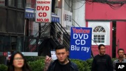 FILE - People walk past a shop selling pirated movie DVDs and music CDs in Beijing, April 27, 2016.