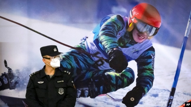 FILE - A police officer wearing a face mask and goggles to protect against COVID-19, stands near a poster of a skier on the wall at a train station in Zhangjiakou in northern China's Hebei Province, Friday, Nov. 26, 2021. (AP Photo/Mark Schiefelbein, File